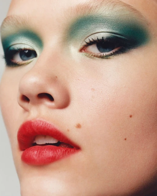 Close-up portrait of an adult female's face featuring prominent green eyeshadow, red lipstick, and clear skin with distinct beauty marks.