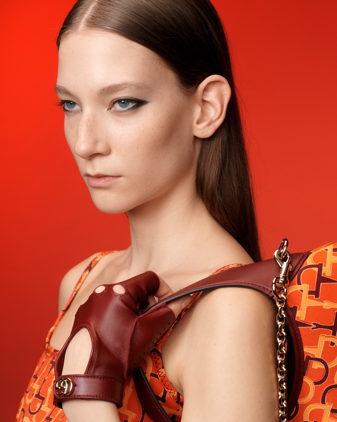 Close-up shot of a fair-skinned adult female model wearing a dress with an orange pattern, burgundy gloves, and a chain-strap purse.