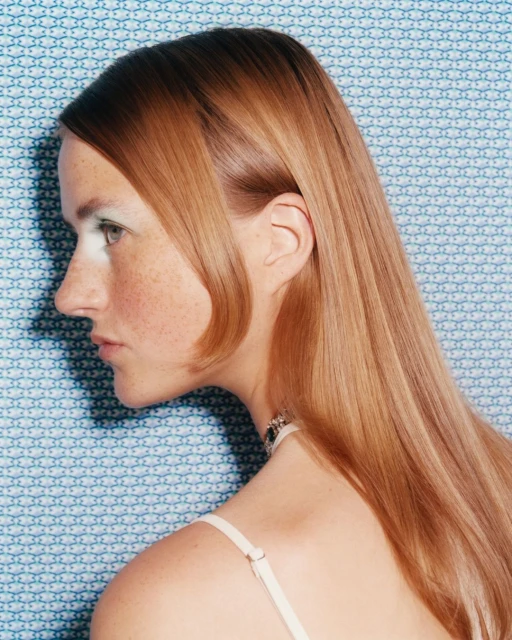 Profile portrait of a woman with long blonde hair wearing a necklace against a blue patterned backdrop, featuring a classic, timeless aesthetic.