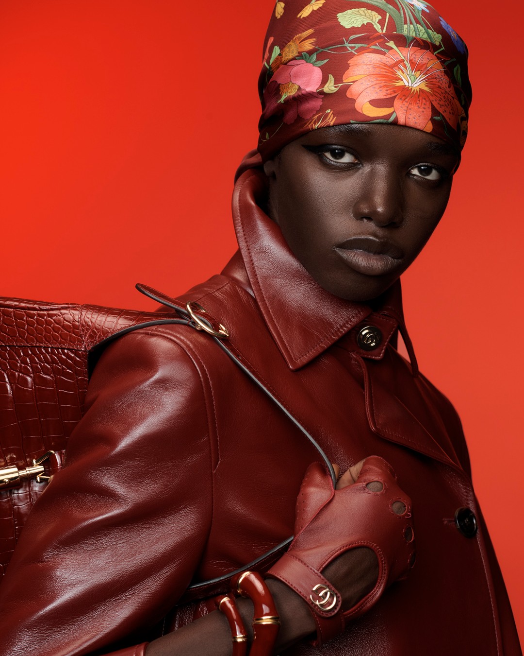 Close-up studio portrait of a Black female model in a patterned headscarf, red leather coat, gloves and accessories against a red background.