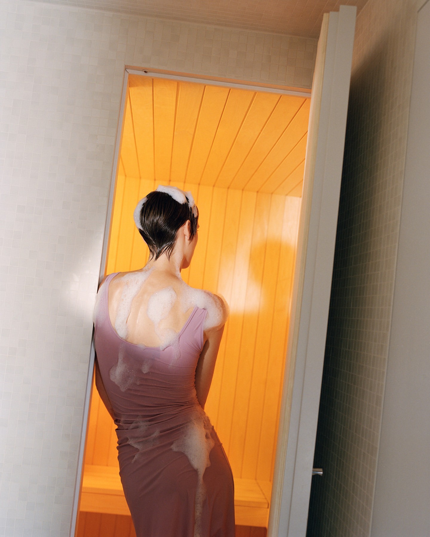 Back view of a slender woman with wet hair in a soaking mauve dress, standing in the open doorway of a hot, bright yellow sauna.