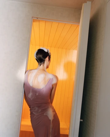 Back view of a slender woman with wet hair in a soaking mauve dress, standing in the open doorway of a hot, bright yellow sauna.