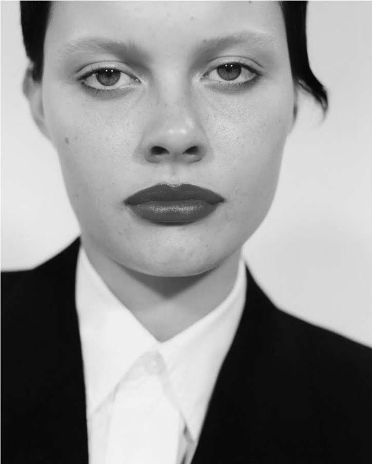 Striking portrait of a young adult woman with pale skin, dark lipstick, wearing a tailored black suit and white shirt, against a clean backdrop.