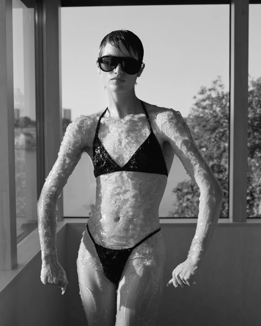 Adult female wearing a black sequin bikini and sunglasses stands indoors against a window with a city view in black and white.