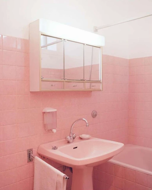 Pink-themed bathroom features a sink, cabinet, mirror, towel, and tiled walls in soft pink hues, creating a retro and clean aesthetic.