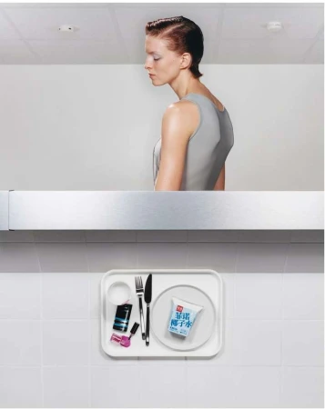 Woman in grey top stands near a tray with drink, cosmetics, and utensils in a bright, minimalist indoor setting. Studio shot.