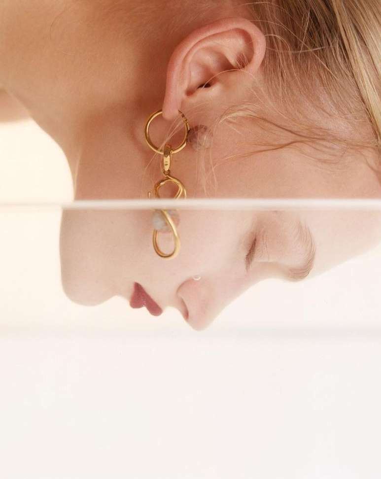 Adult female wearing gold statement earring with her face reflected in a glass pane on a light-peach backdrop in an indoor setting.
