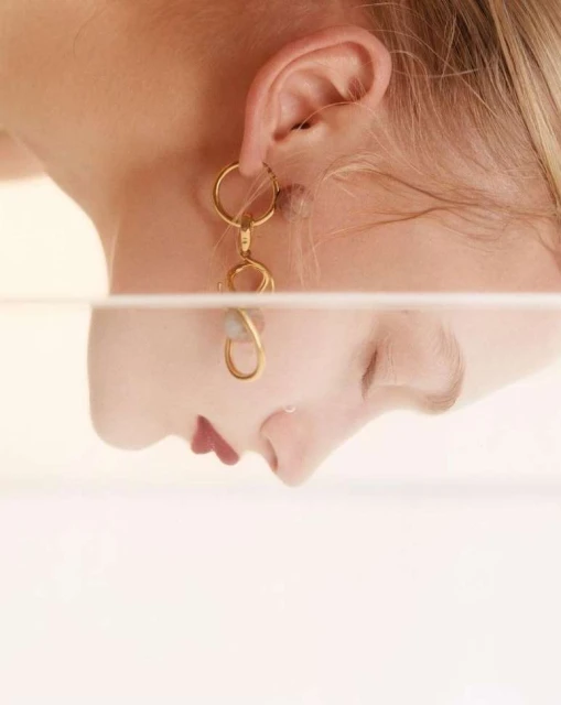 Adult female wearing gold statement earring with her face reflected in a glass pane on a light-peach backdrop in an indoor setting.