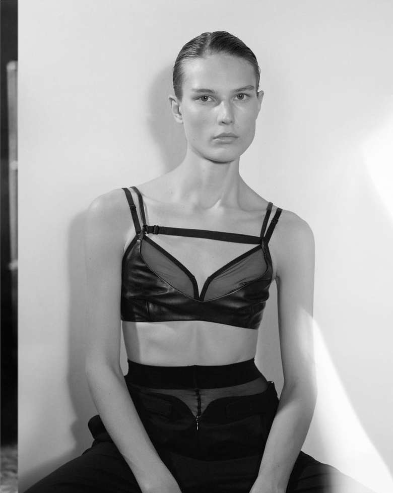 Black and white portrait of a model wearing a black bra top and pants, posing in front of a white backdrop with natural light.