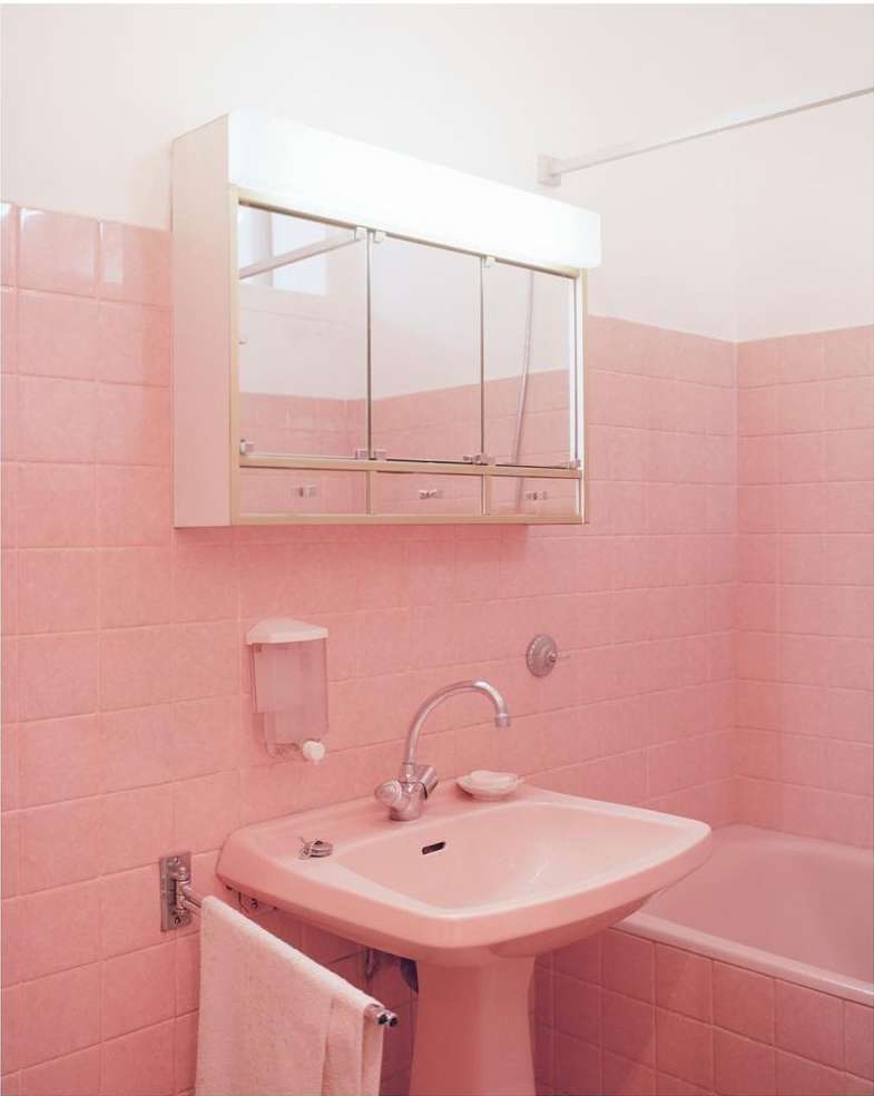 Bathroom interior with pink tiled walls, a pedestal sink, mirrored cabinet, and white towel. The scene is brightly lit with a clean, simple aesthetic.