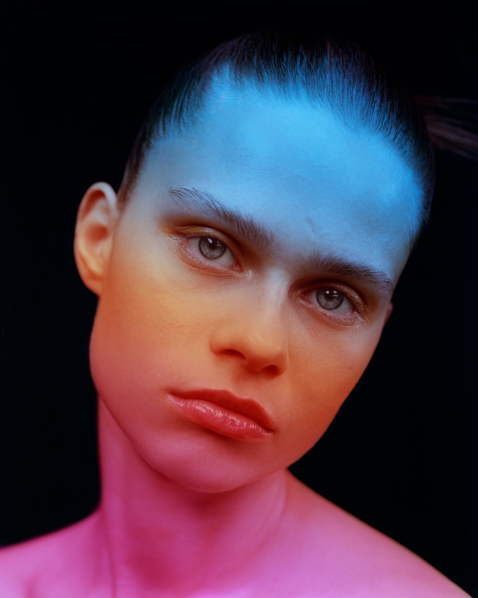 Close-up studio portrait of an adult female model with slicked-back hair, illuminated with pink and blue lighting against a black backdrop.