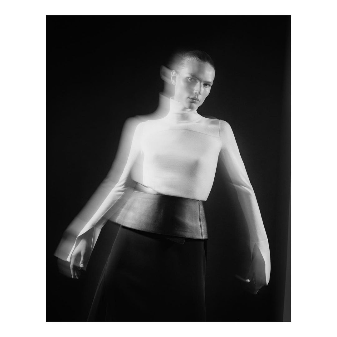 Black and white fashion photograph of a woman wearing a white turtleneck and black skirt, captured with motion blur effect, set against a dark backdrop.