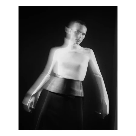 Black and white fashion photograph of a woman wearing a white turtleneck and black skirt, captured with motion blur effect, set against a dark backdrop.