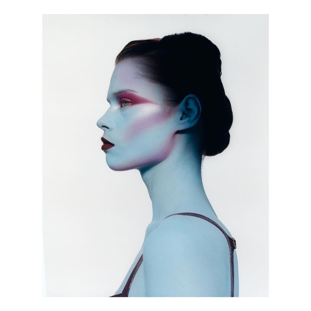 Profile portrait of a young woman with blue skin and bold pink makeup against a white background, showcasing beauty and fashion.