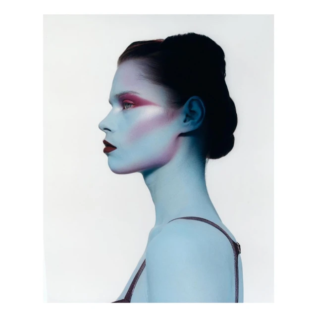 Profile portrait of a young woman with blue skin and bold pink makeup against a white background, showcasing beauty and fashion.