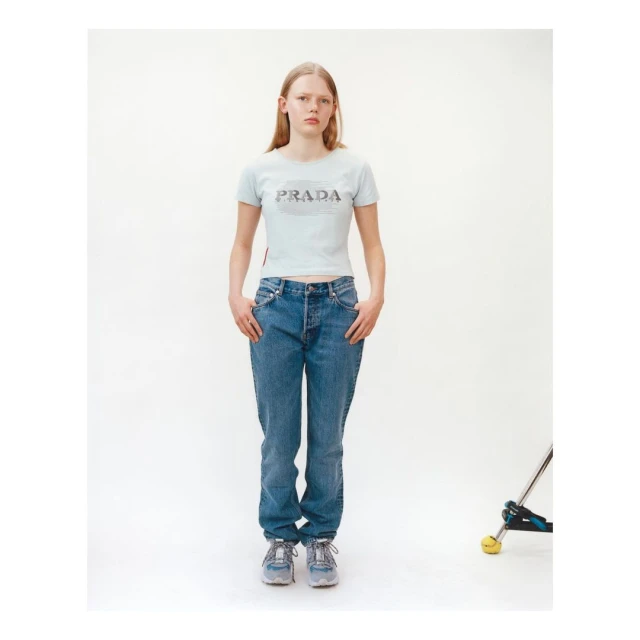 A fair-skinned young woman in a Prada t-shirt and blue jeans stands in front of a white background in a fashion-style studio photograph.