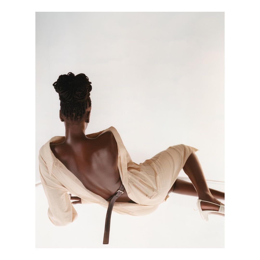 Rear view of a woman with updo hairstyle lying on a reflective surface, wearing a beige dress, brown belt, and white heels, against a white backdrop.