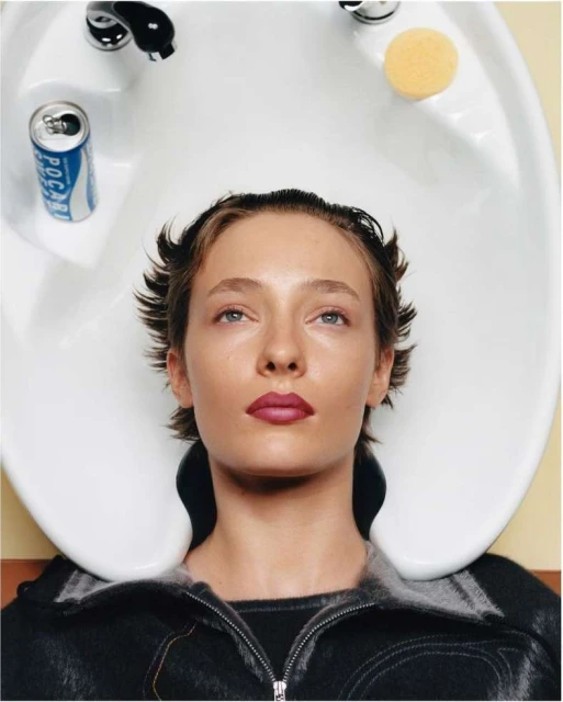 Adult woman with short brown hair leans back into a white salon washing basin with a can of soda, faucet, and yellow sponge visible.