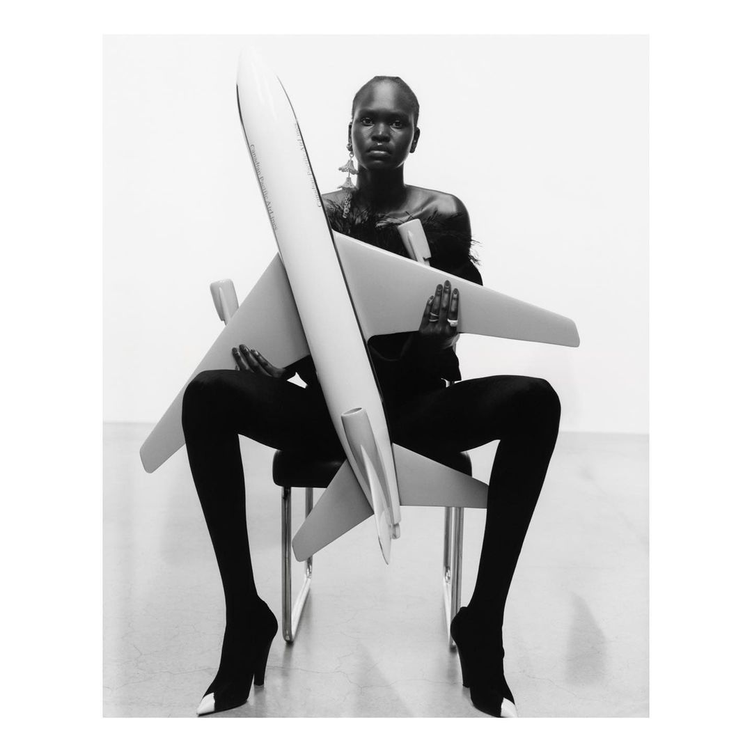 Black and white studio portrait of an adult woman holding a toy airplane while seated, wearing dark clothing and high heel shoes.