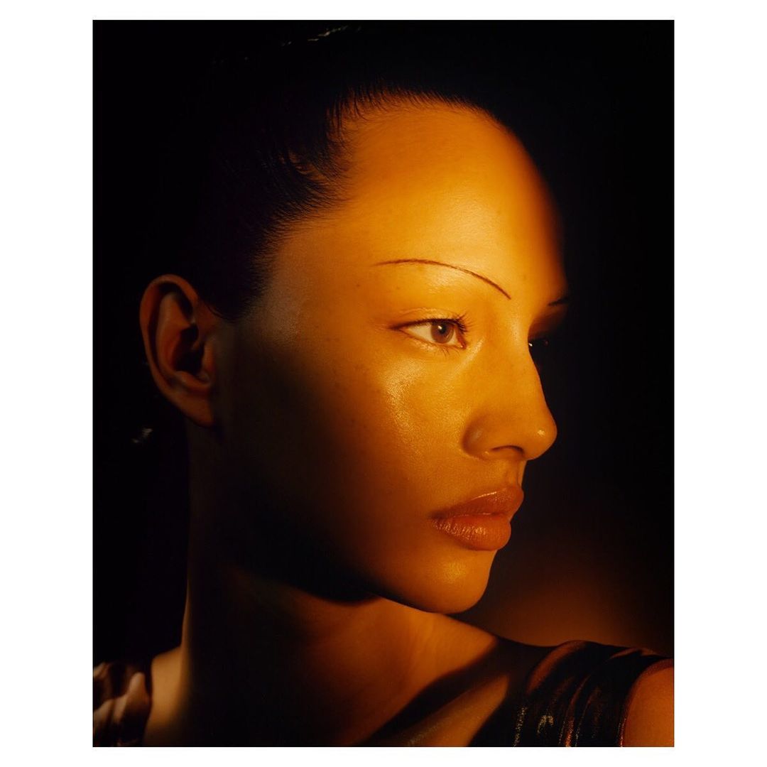 Close-up portrait of an adult woman with dark hair and thin eyebrows, featuring side lighting, a shallow depth of field, and a dark background.