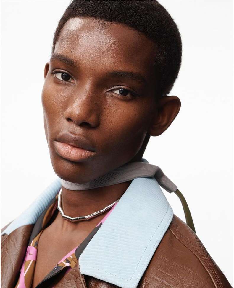 Close-up shot of a dark-skinned male model wearing a silver necklace and brown leather jacket in a bright, high-key studio setting.