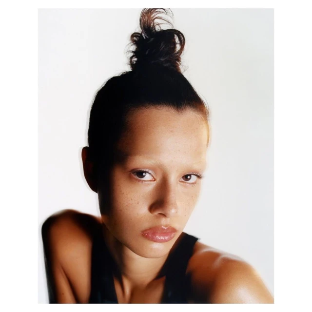 Close-up portrait of a young adult woman with dark hair styled in a bun, light skin, and light eyebrows against a bright white background.