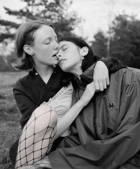 Monochrome portrait of two young women embracing outdoors, one in fishnet stockings and Kappa jacket, resting against each other.
