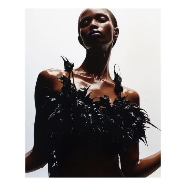 Glamourous portrait of a black woman with slicked-back hair and purple lipstick wearing black feathered top in a white studio.