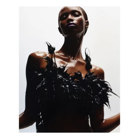 Glamourous portrait of a black woman with slicked-back hair and purple lipstick wearing black feathered top in a white studio.