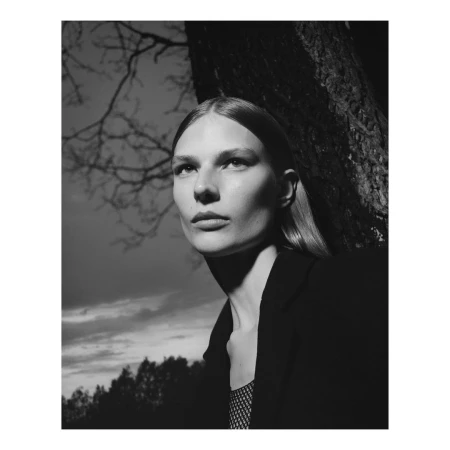 Monochrome portrait of a female model standing outdoors leaning against a tree trunk under a cloudy sky.
