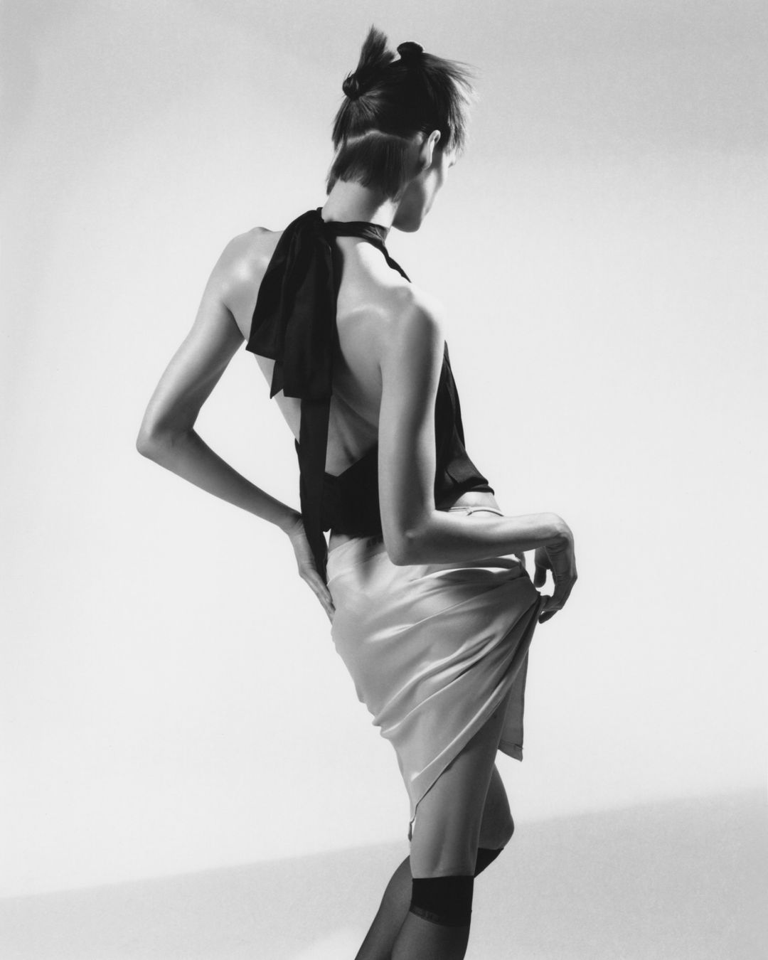 Fashion shot of a slender, fair-skinned woman in a black top and light skirt, set against a plain white background in monochrome.