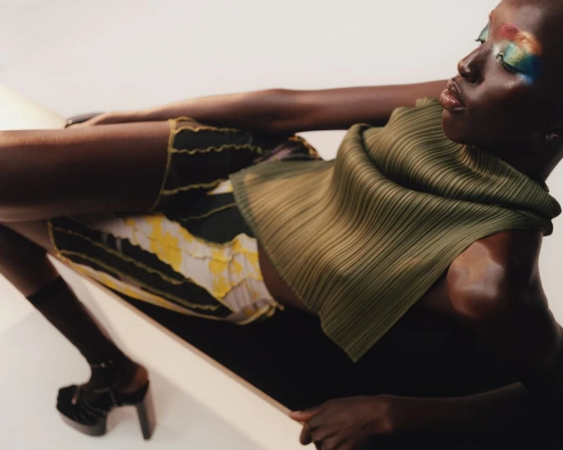 Fashion shot of a Black model with colorful eye makeup, wearing an olive green top and patterned shorts in a minimalist indoor setting.