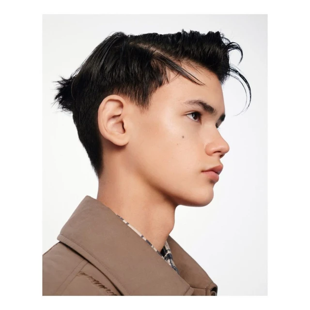 Profile portrait of a young adult male with dark, styled hair wearing a neutral trench coat against a plain white background.