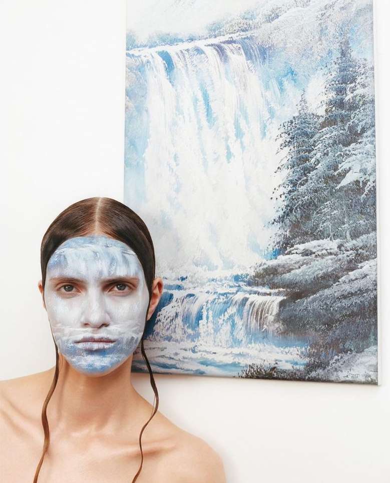 Woman's face with painted waterfall design, portrait-style, with a waterfall painting in background, studio lighting and indoor setting.