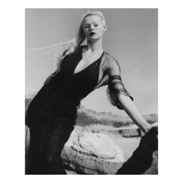 Black and white portrait of a woman in a dark dress posing on rocks in a desert landscape, facing the camera with a neutral expression.