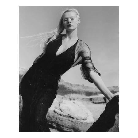 Black and white portrait of a woman in a dark dress posing on rocks in a desert landscape, facing the camera with a neutral expression.