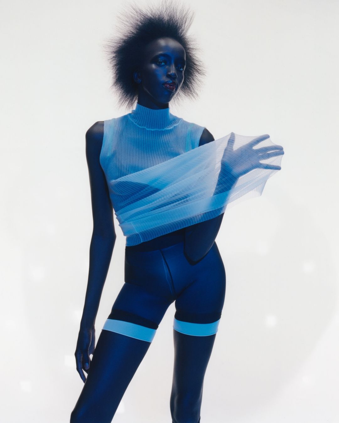 A fashion shot of a Black female model in a studio with a blue tint, wearing a sleeveless top, shorts, and holding a sheer fabric.