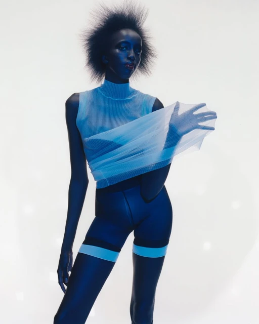 A fashion shot of a Black female model in a studio with a blue tint, wearing a sleeveless top, shorts, and holding a sheer fabric.
