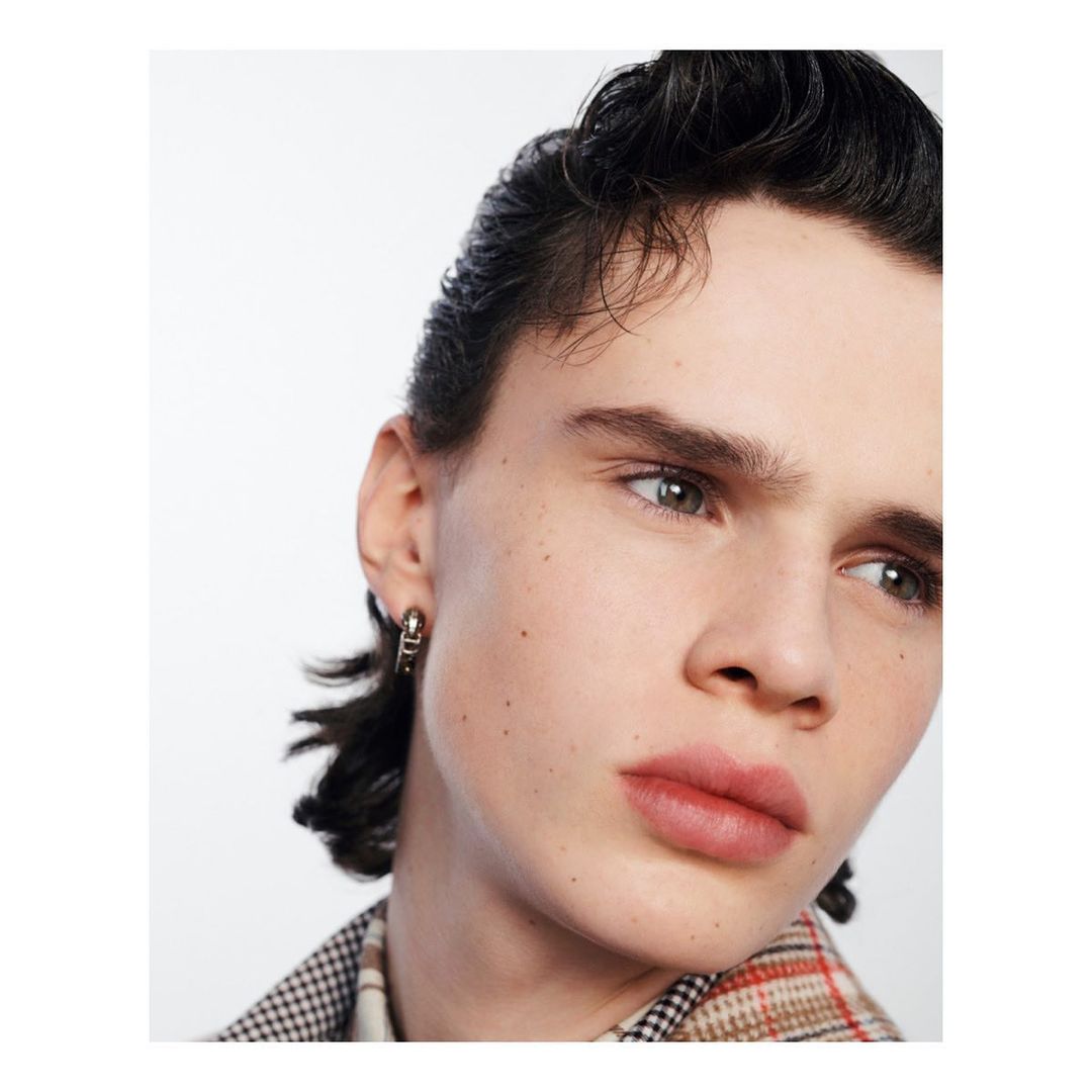 Close-up portrait of a young man wearing a plaid patterned shirt and a small metallic hoop earring against a plain white background.