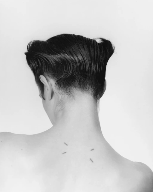 Monochrome shot of a person with dark, wavy hair, bare back, and minimalist tattoo against a plain white background, captured in a studio setting.