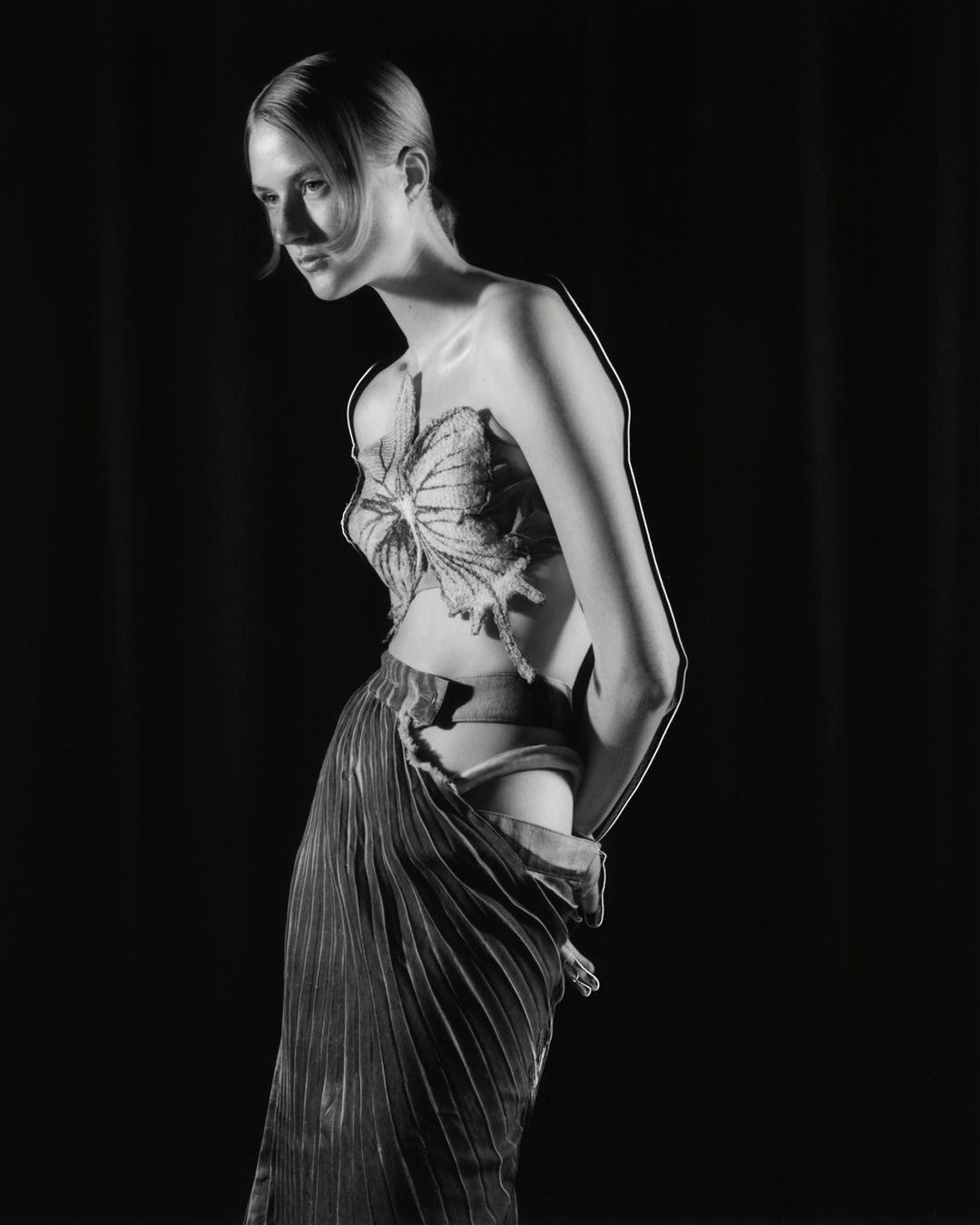 Monochrome studio portrait of a slender woman in an ornate, butterfly-like top and pleated skirt, against a black backdrop. 