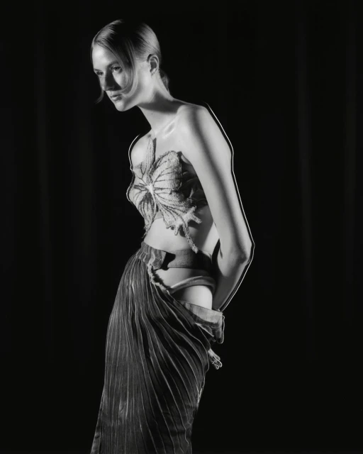 Monochrome studio portrait of a slender woman in an ornate, butterfly-like top and pleated skirt, against a black backdrop. 