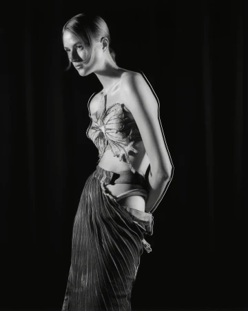 Monochrome studio portrait of a slender woman in an ornate, butterfly-like top and pleated skirt, against a black backdrop. 