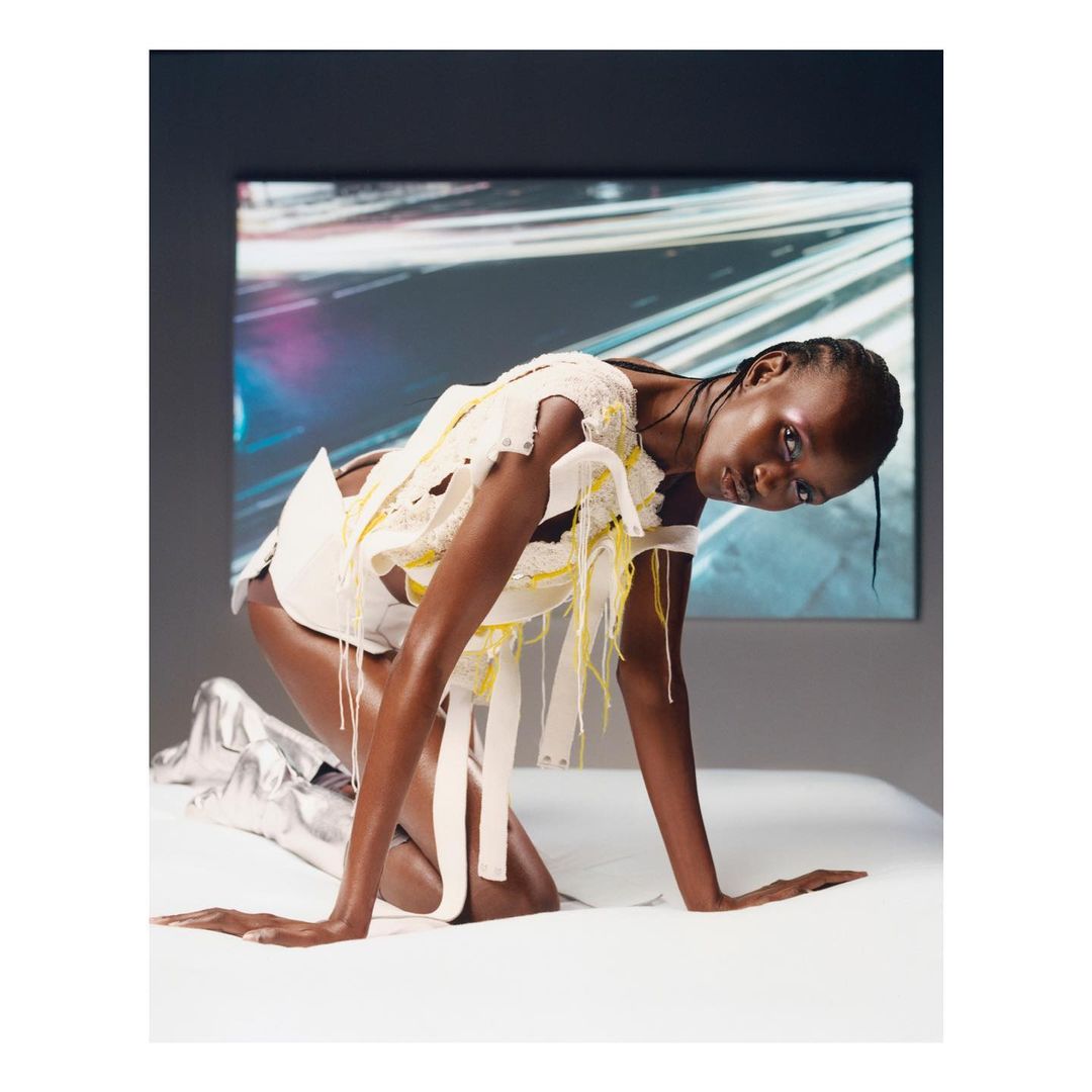 Woman with dark skin kneels wearing ripped white avant-garde clothing in a photo studio against a blurred color display screen.