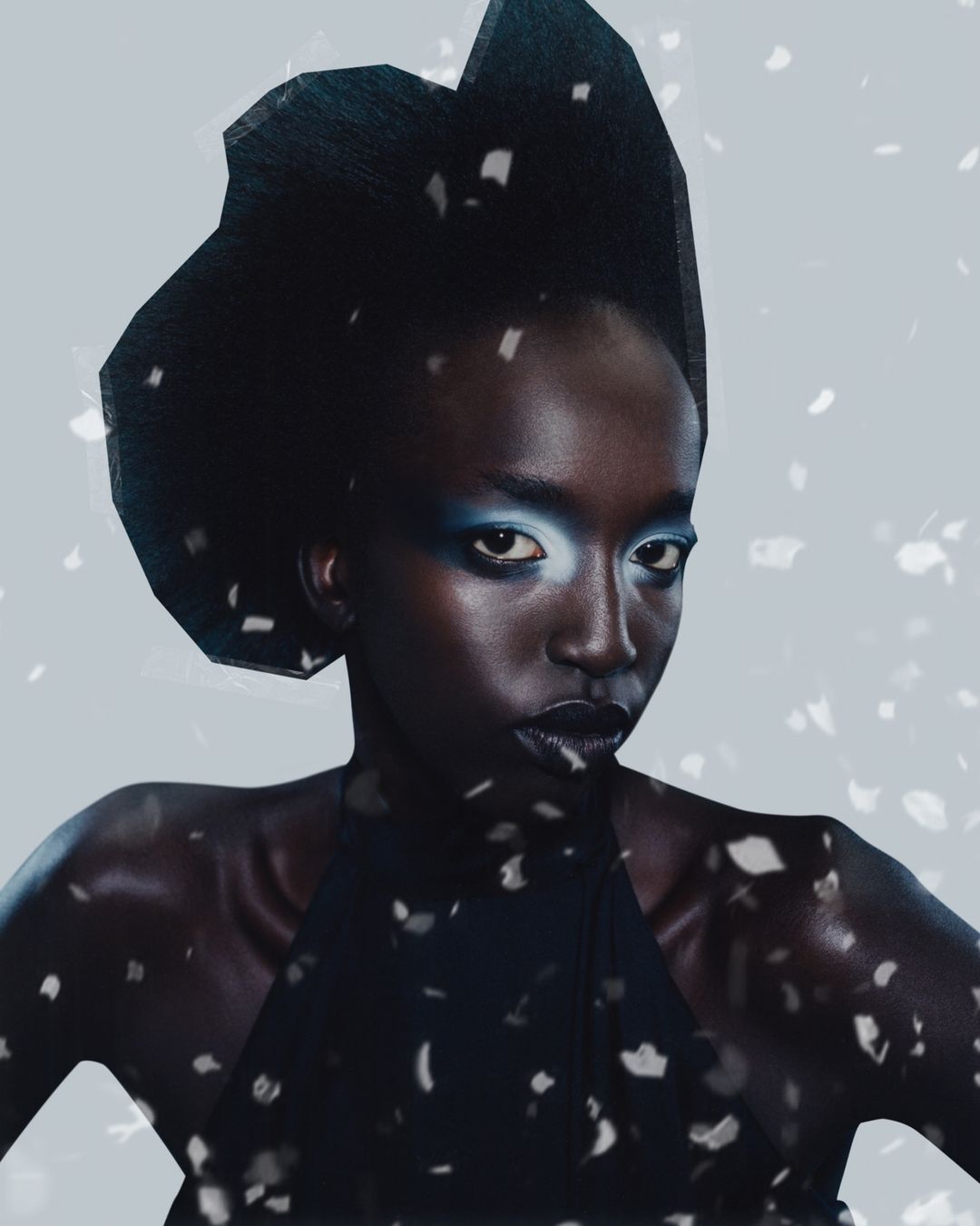 Dark-skinned woman with blue makeup and a black headpiece poses indoors with a light background and scattered white elements.