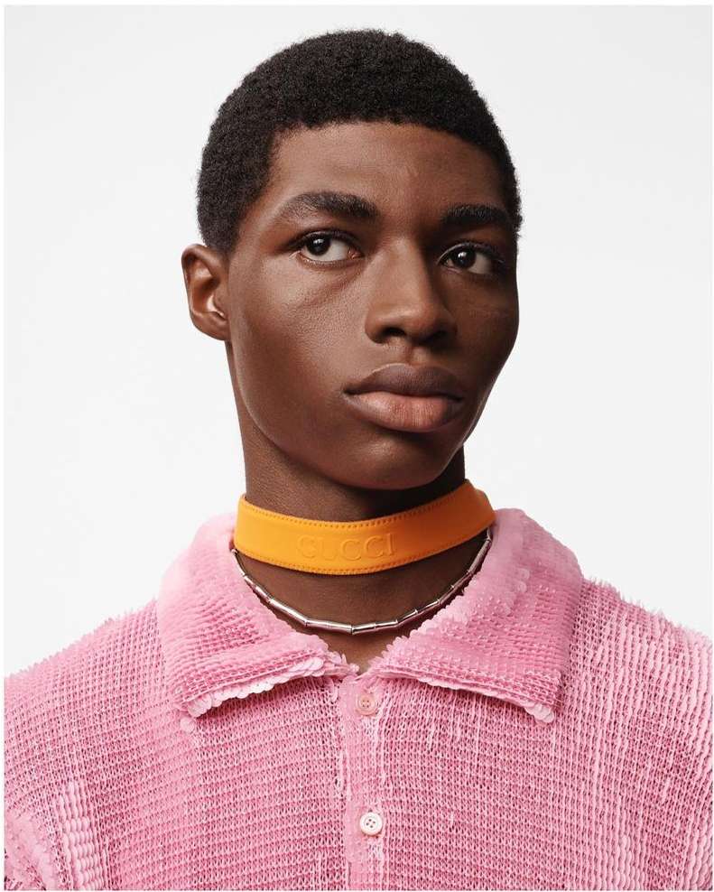 Close-up portrait of a dark-skinned male model wearing an orange choker and pink shirt against a white backdrop in a studio.