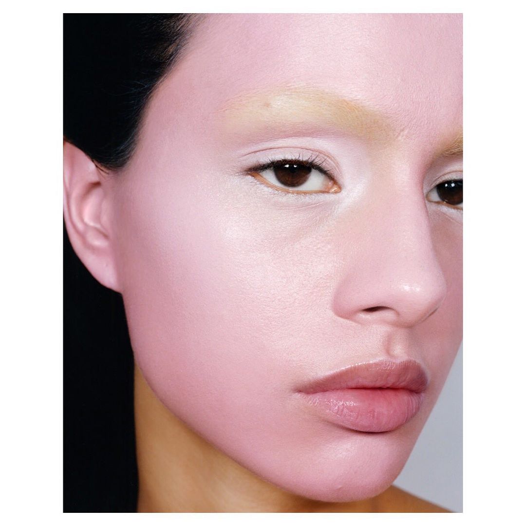Close-up beauty shot of a woman with light pink makeup, light eyebrows, brown eyes and dark hair against a light gray background.