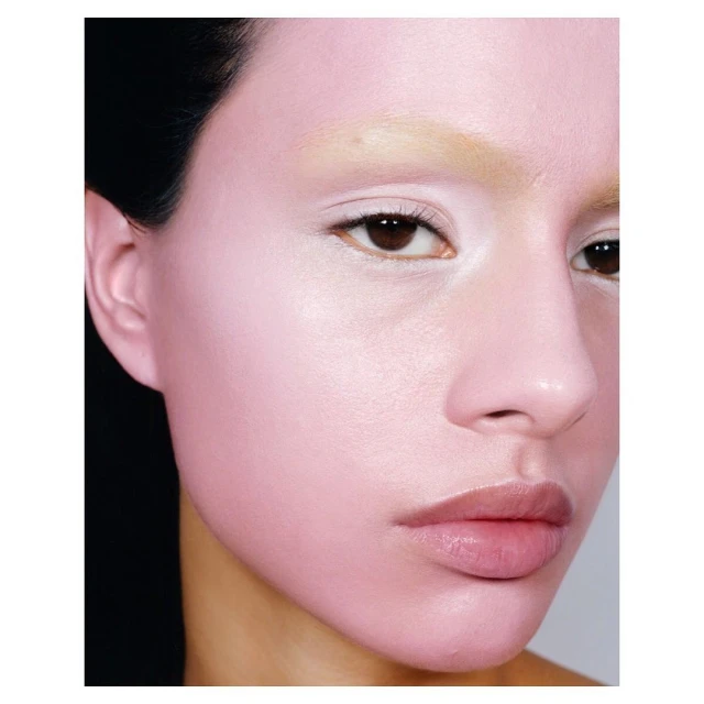Close-up beauty shot of a woman with light pink makeup, light eyebrows, brown eyes and dark hair against a light gray background.