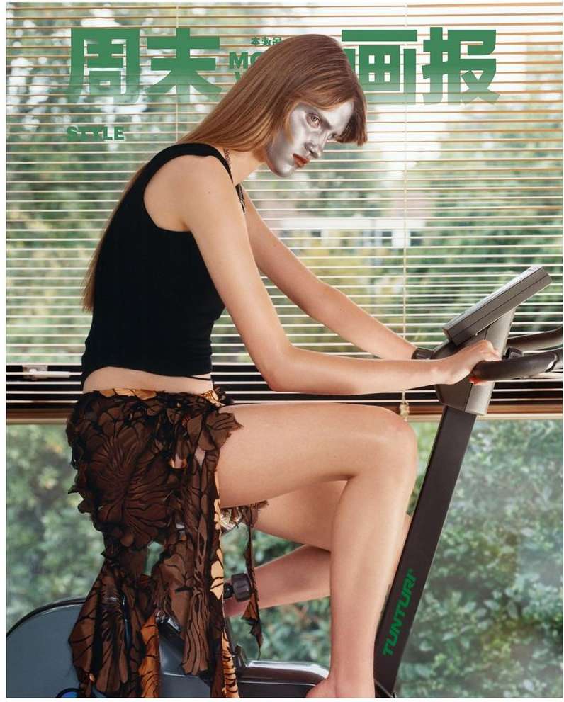 Adult female on exercise bike with metallic face makeup in front of blinds, styled for a fashion magazine cover, indoors.