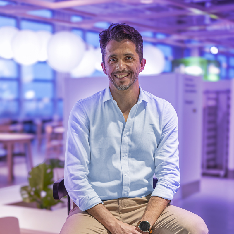 A smiling adult man in a light blue shirt is sitting on a chair in an indoor space with bright lighting and tables and chairs in the background.
